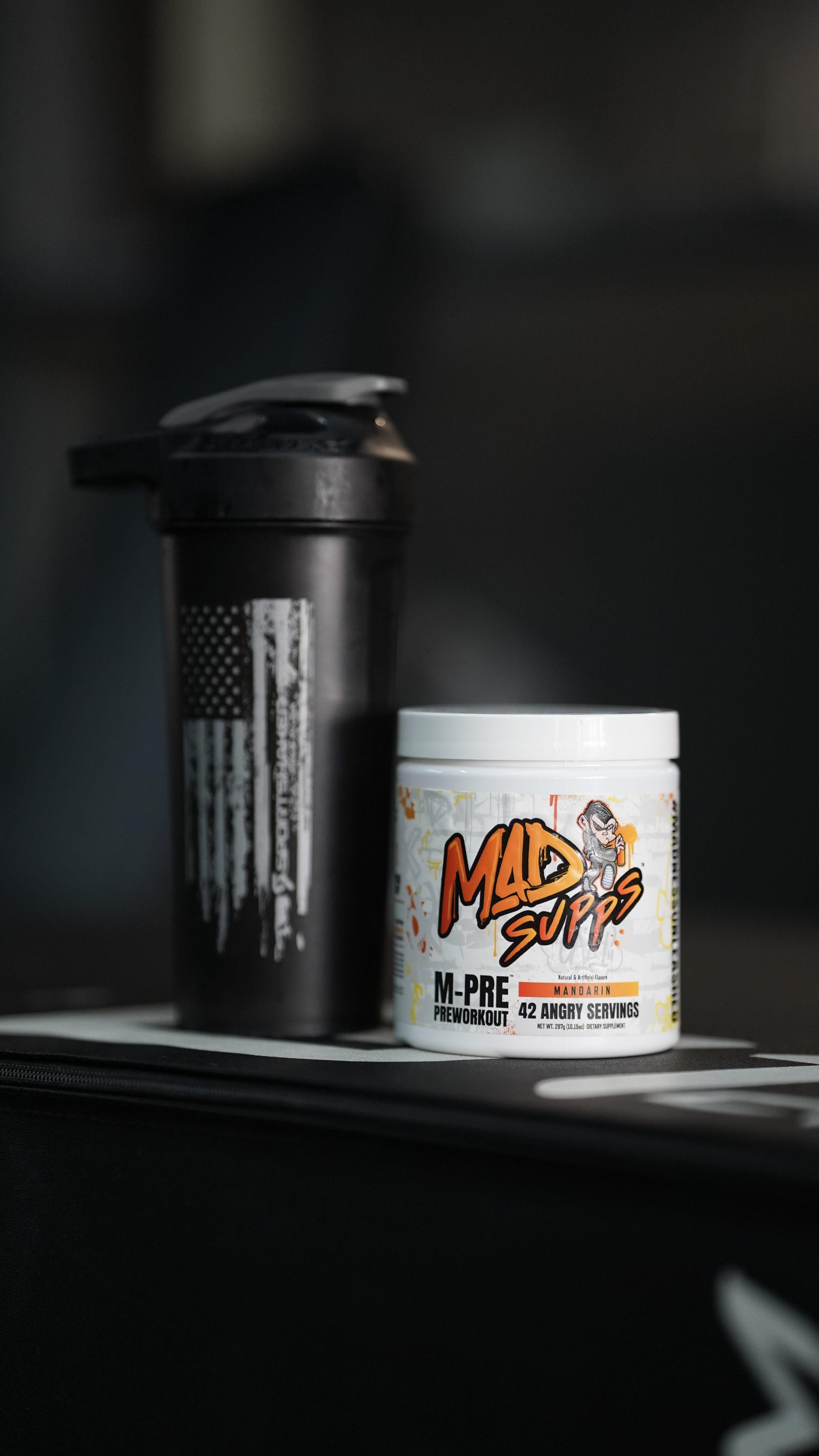 A black protein shaker bottle beside a white container labeled MAD PUPS M-PRE preworkout mandarins on a dark surface.