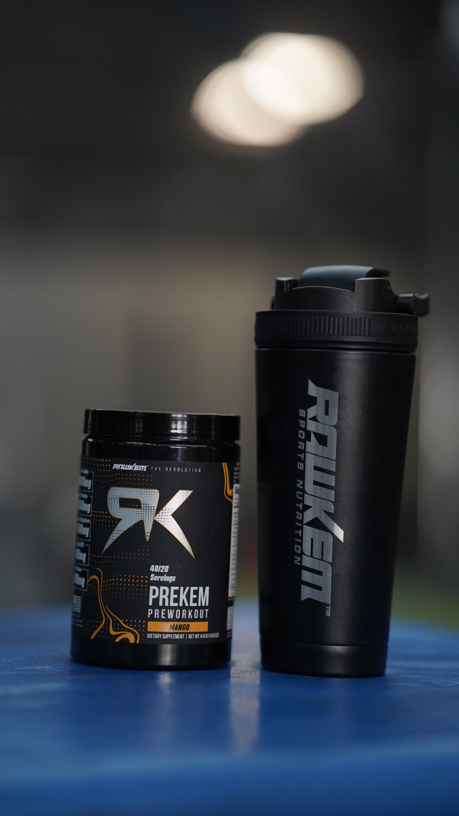 Two black fitness containers on a blue surface: a jar labeled Prekem Preworkout Mango and a tall black shaker bottle beside it, set against a blurred background.