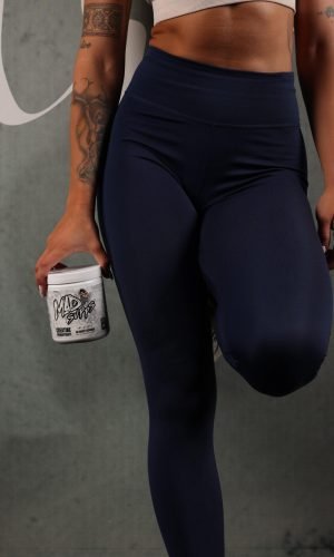 Person wearing navy leggings holding a protein shaker, tattoos visible on the arm, against a neutral backdrop.