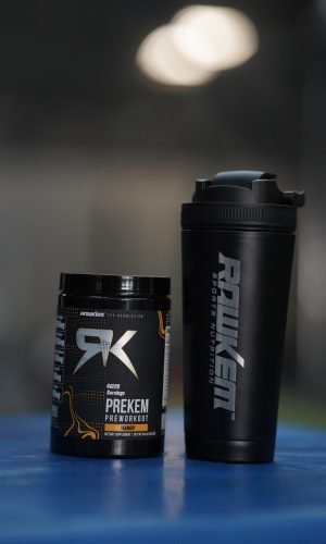 Two black fitness containers on a blue surface: a jar labeled Prekem Preworkout Mango and a tall black shaker bottle beside it, set against a blurred background.